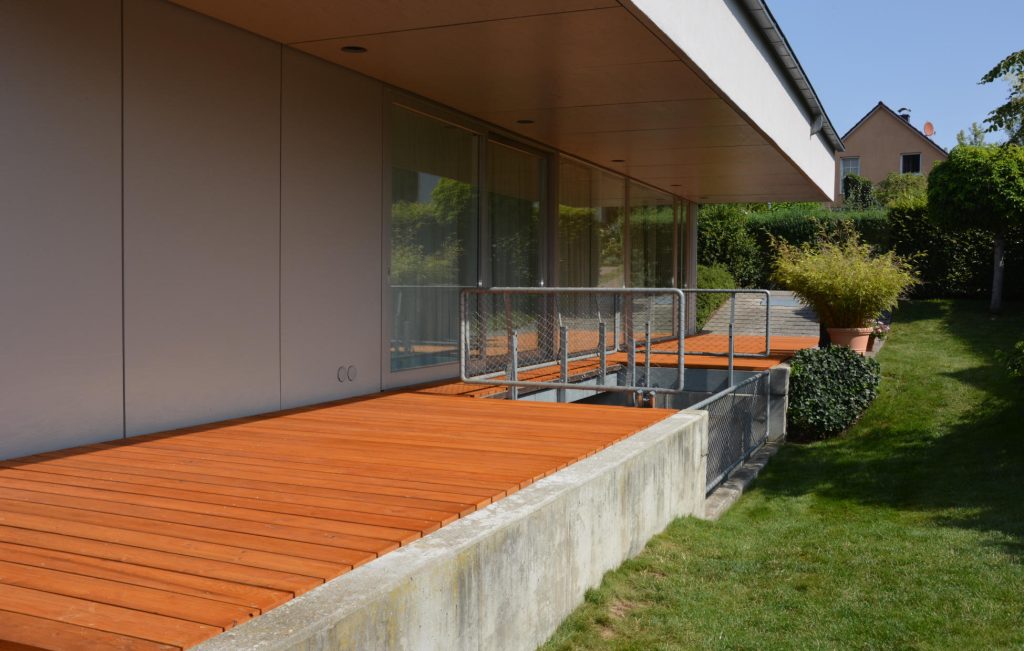 Holzdeck an der Seite eines Hauses, umgeben von Grün, ideal für Entspannung und gesellige Zusammenkünfte. Terrassenplanung
